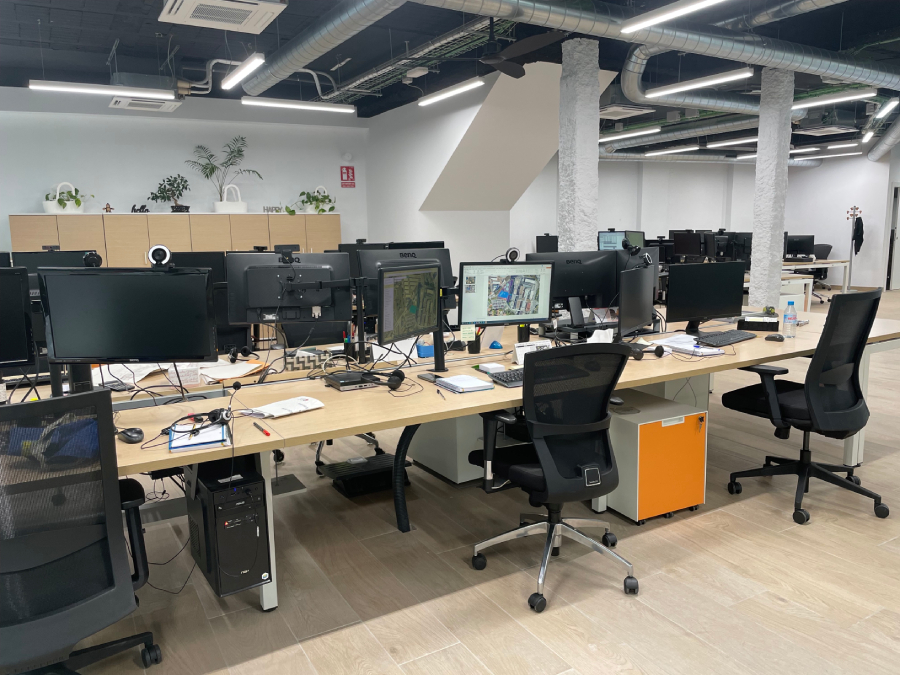 Un moderno espacio de oficina con escritorios equipados con monitores, sillas ergonómicas, techo oscuro con conductos expuestos e iluminación moderna.