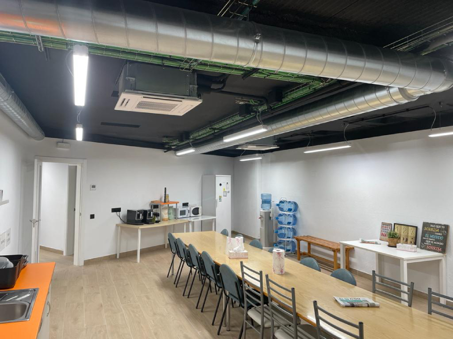 Área de comedor o descanso de oficina con una larga mesa de madera, zona de cocina, dispensador de agua y techo con conductos expuestos.