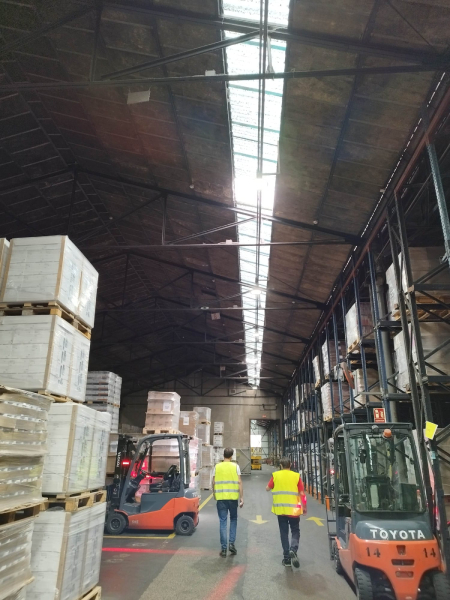 Interior de un almacén industrial con estanterías altas, montacargas Toyota en operación y dos trabajadores con chalecos reflectantes caminando por el pasillo principal.